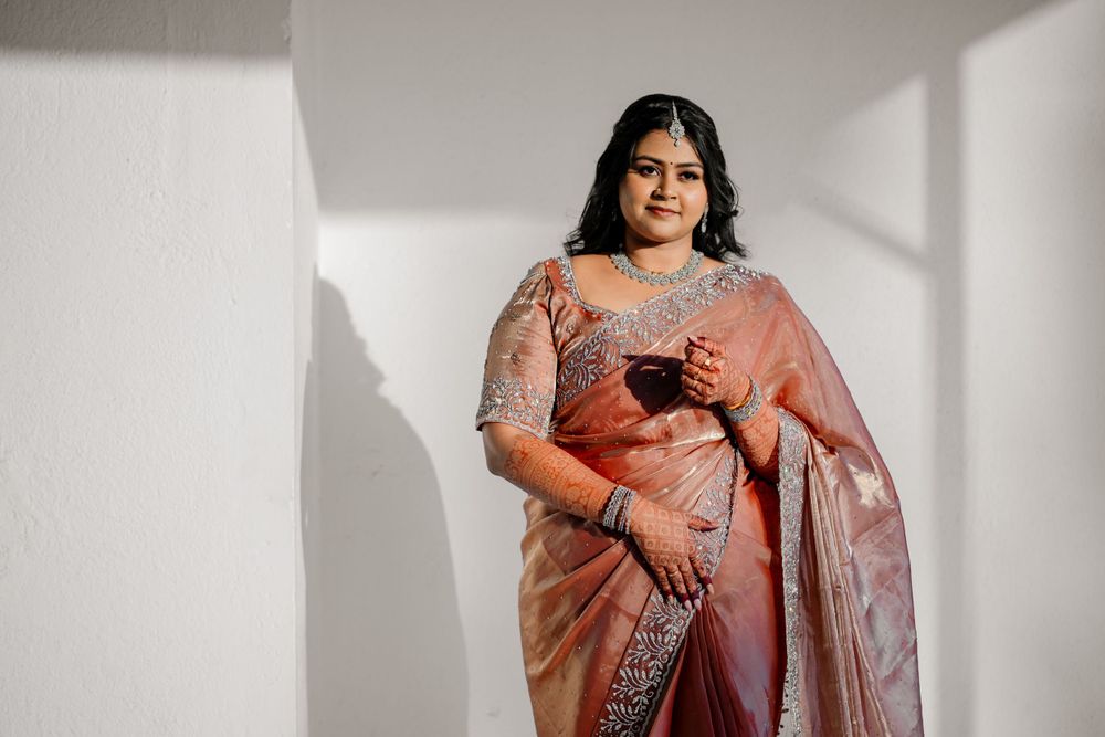 A curvy woman in an embellished peach-colored saree poses elegantly against a white wall with dramatic shadows.