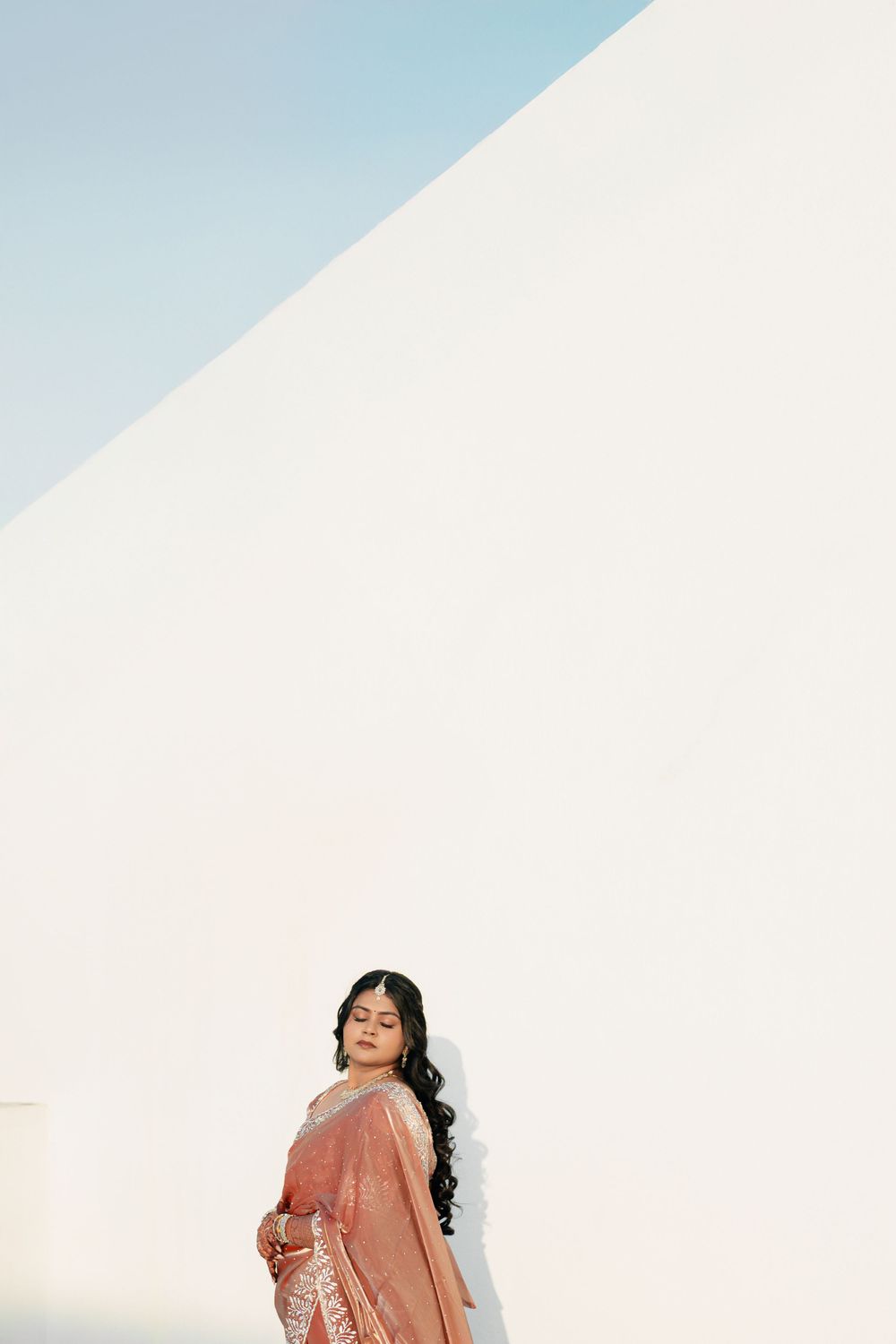 Minimalist portrait of bride in peach saree against white architectural background.