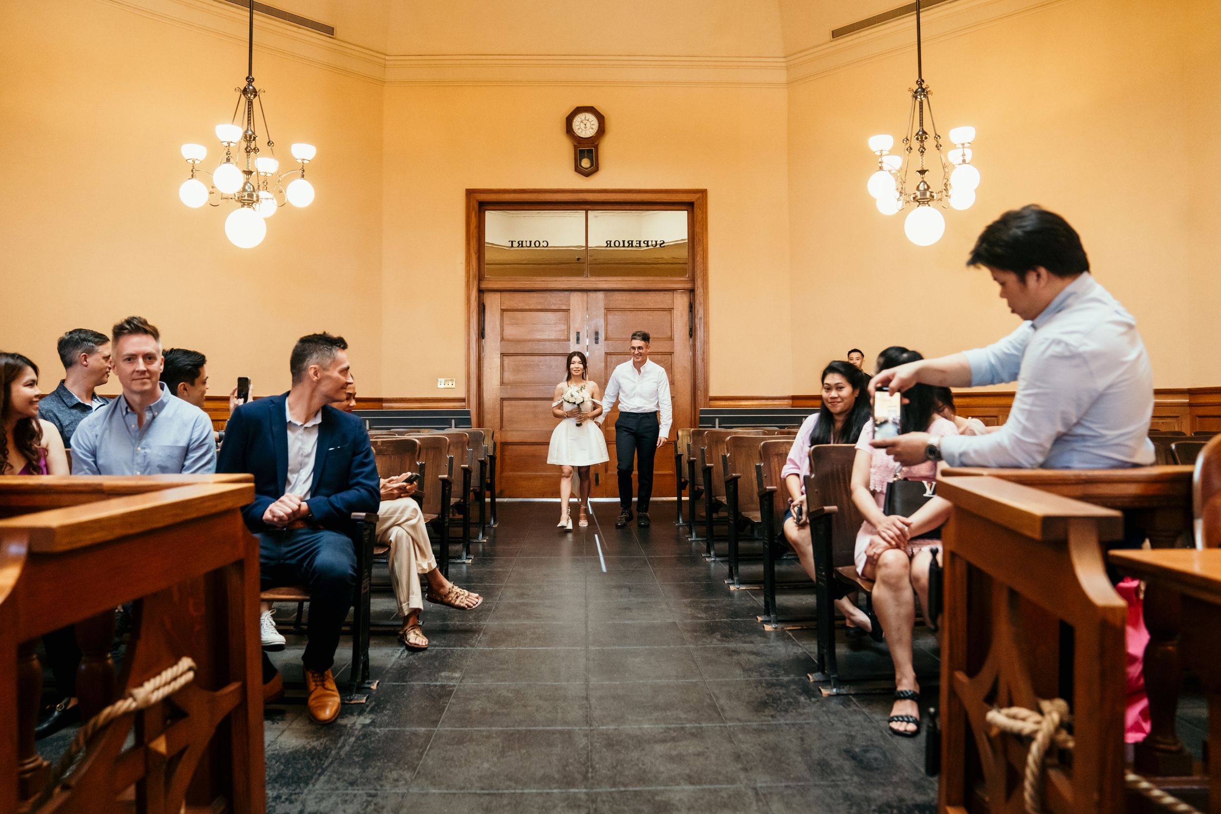 People gather inside a historic courthouse room with wooden pews, pendant lights, and a doorway at one end.