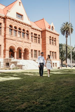 A couple walks on a lawn in front of a historic red brick building with Spanish architectural features and a tall palm tree.