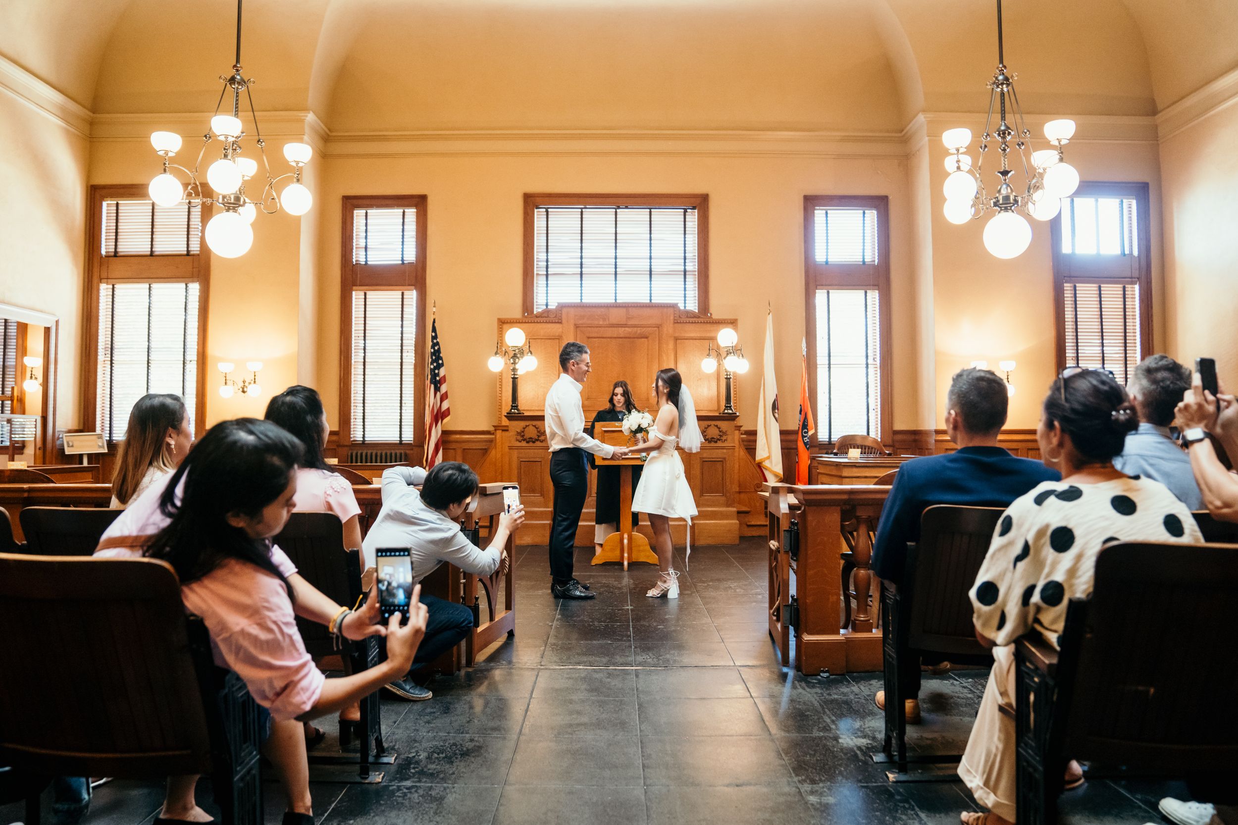 People seated on wooden pews inside a historic courtroom with warm lighting and tall windows during a legal proceeding.