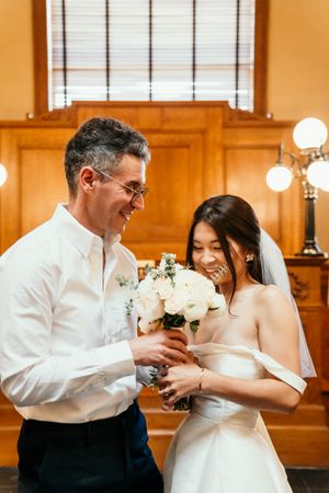 A bride in a white off-shoulder dress shares a joyful moment with her partner while holding a white floral bouquet.