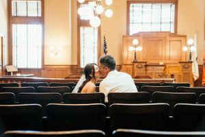 Two people share an intimate moment while sitting in wooden pews of an empty courtroom illuminated by warm lighting.