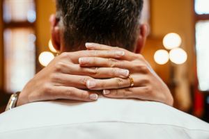 Hands clasped behind neck showing gold wedding ring with soft bokeh lighting in background.