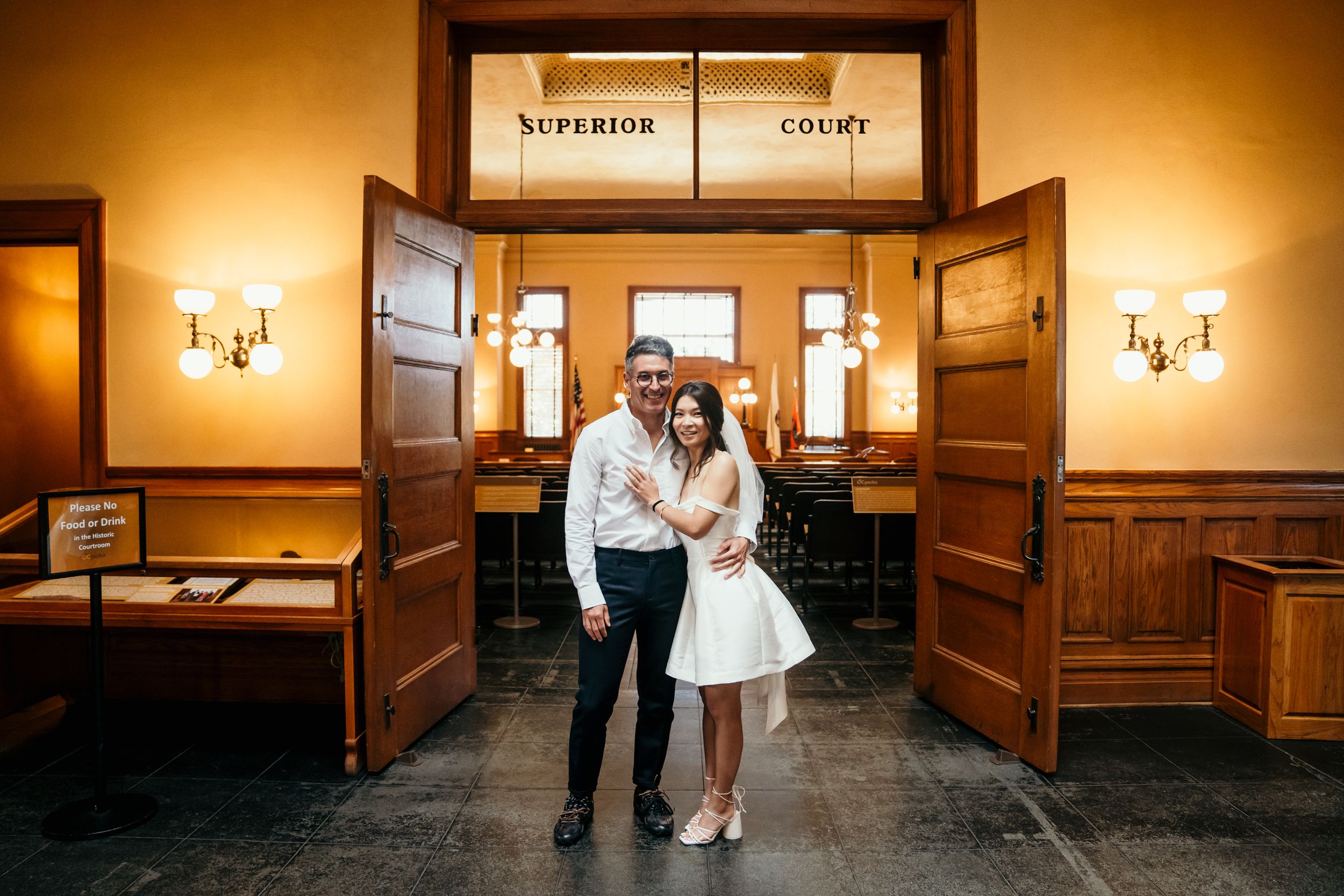 A couple embraces in front of wooden courthouse doors with warm ambient lighting and vintage wall sconces.