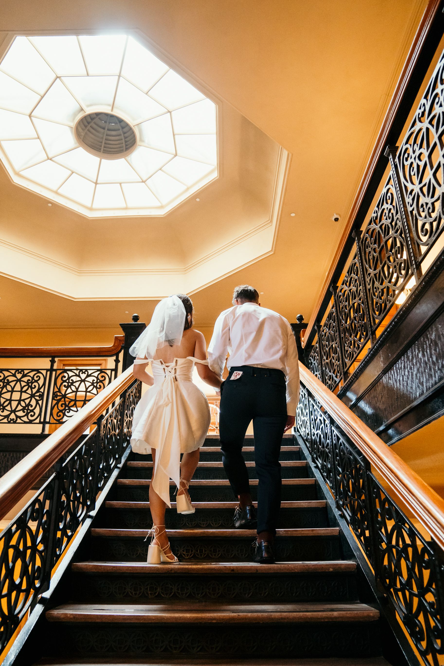 A couple ascends an elegant staircase with ornate iron railings beneath a backlit octagonal skylight in a luxury venue.