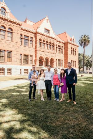 A group poses in front of a historic pink stone building with arched windows and palm trees.