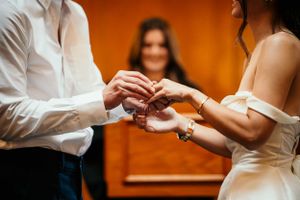 Two people exchanging rings during an intimate wedding ceremony against an orange backdrop.