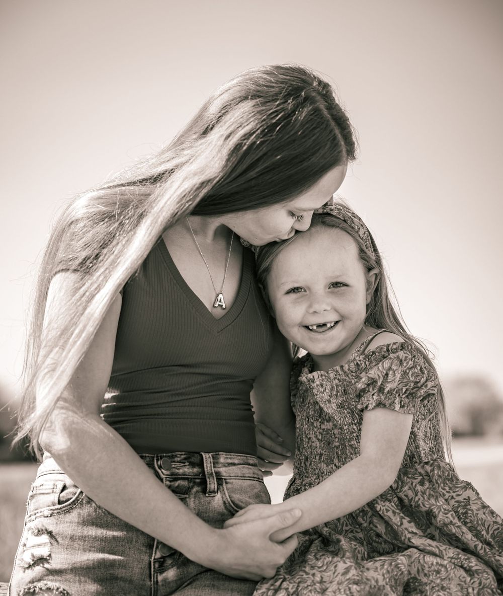 A young child in a floral dress hugging someone affectionately outdoors in a black and white portrait.
