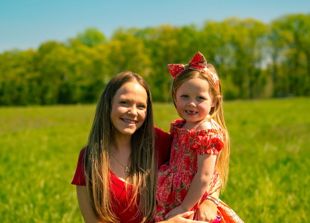 A parent and child wearing matching red outfits pose together in a sunny meadow with green trees in the background.