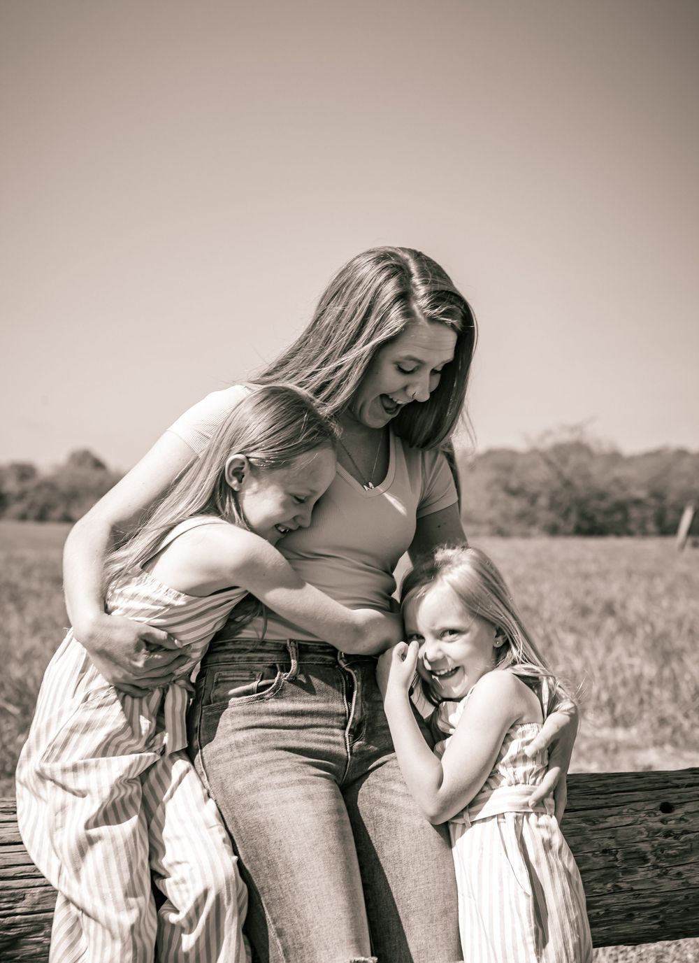 A tender family moment in sepia tones as two children in matching dresses hug their mother on a sunny day outdoors.