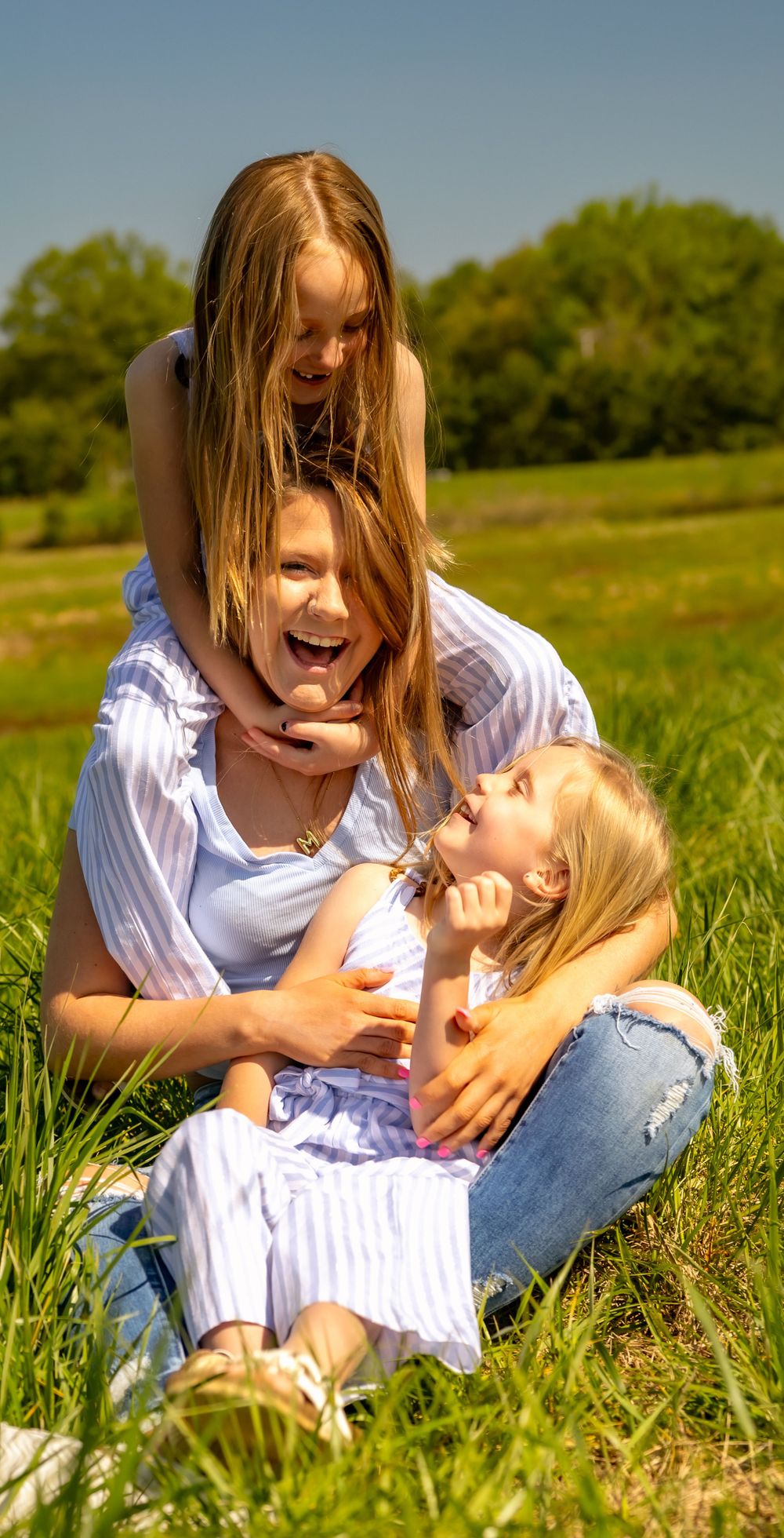 Three people with blonde hair share joyful laughter while relaxing together in a sunny green meadow on a summer day.