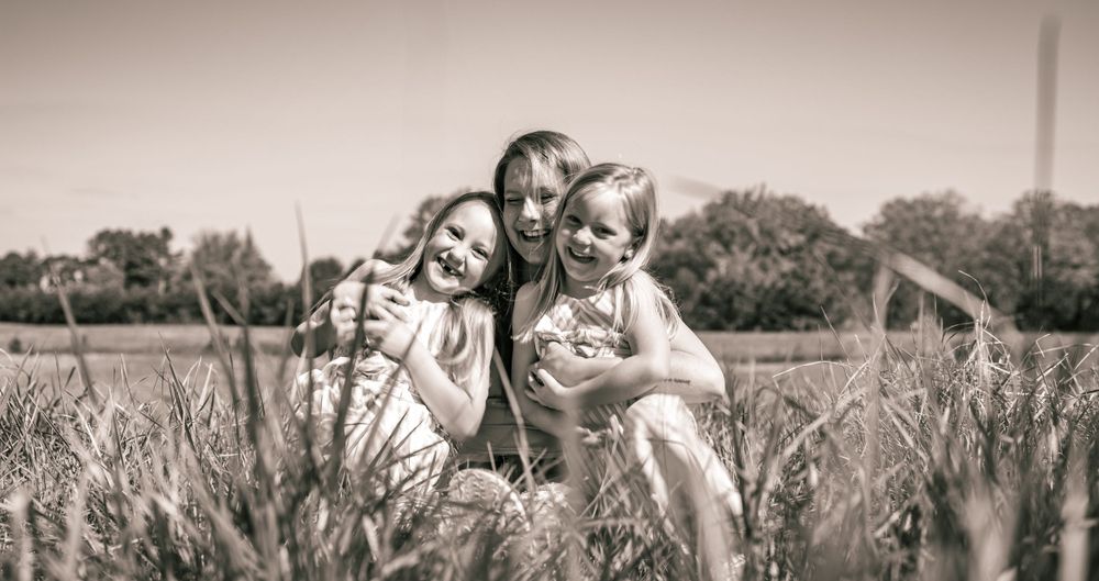 A sepia-toned scene of three people sitting together in a grassy field surrounded by tall plants and trees.