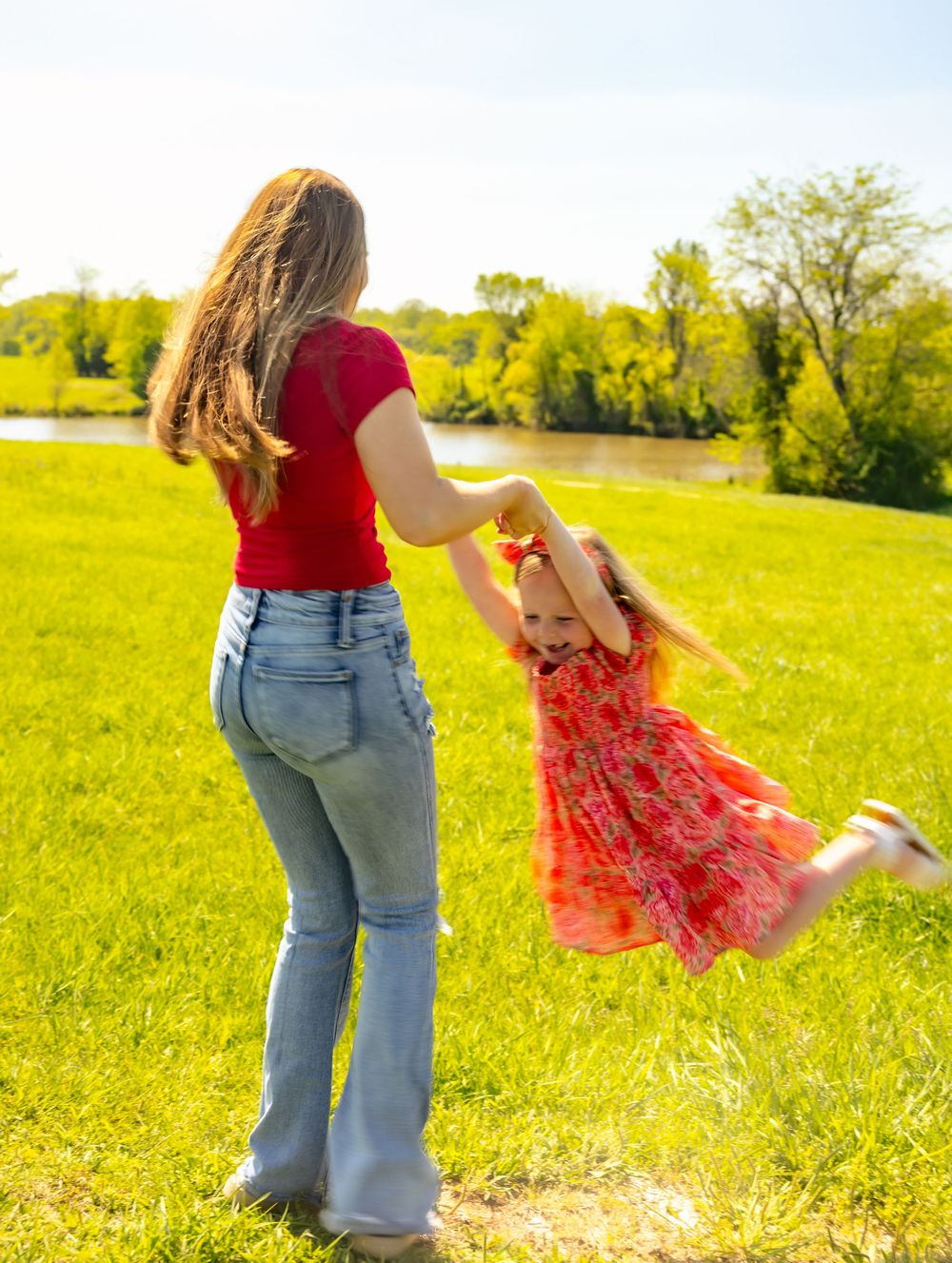 A person spins a companion in a red dress through a sunny meadow near a lake.
