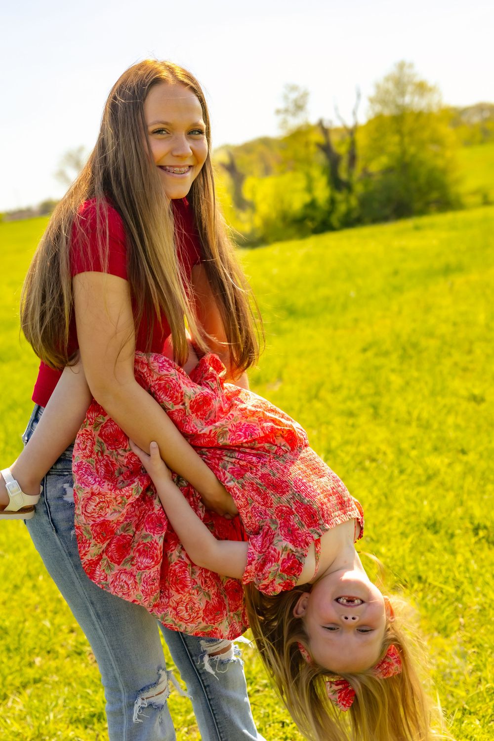 Two people enjoying a playful moment in a sunny meadow with bright yellow flowers and green trees in background.