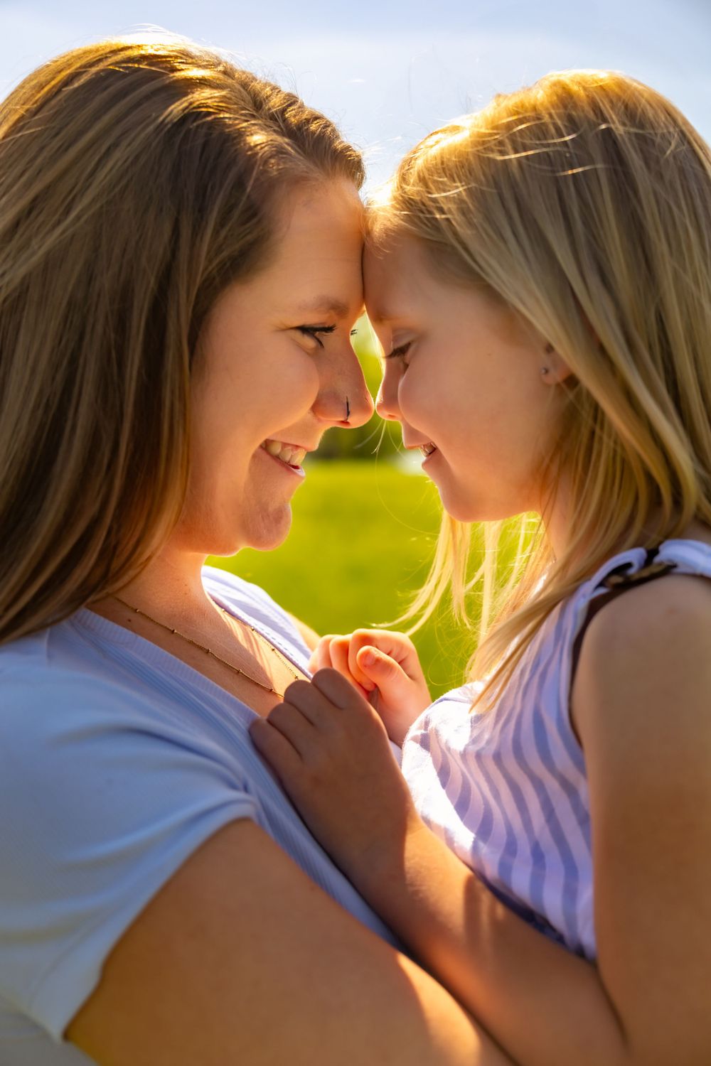 A tender moment between parent and child touching foreheads and smiling in warm sunlight outdoors.
