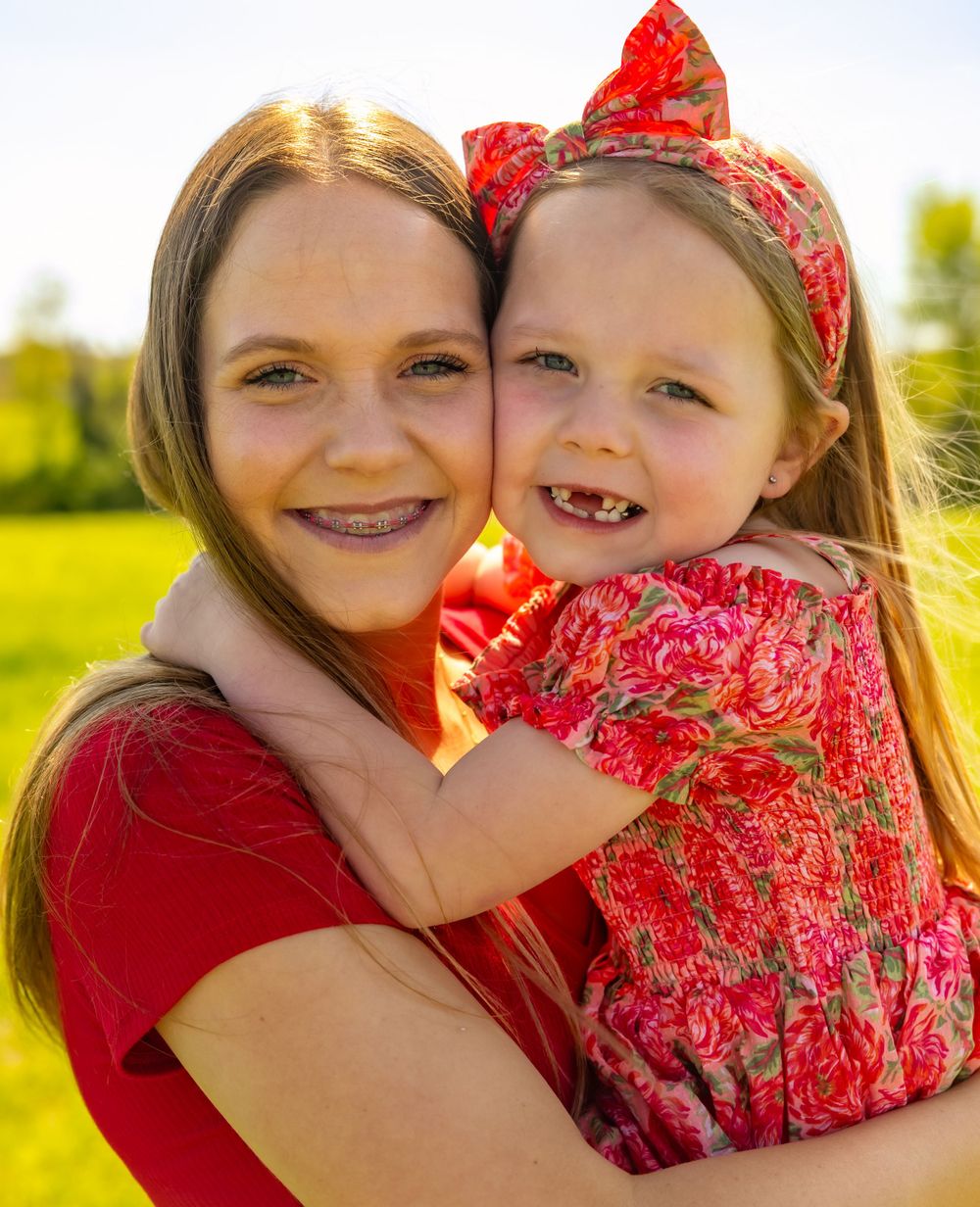 A parent and child wearing matching red outfits embrace while smiling on a sunny day in a grassy field.