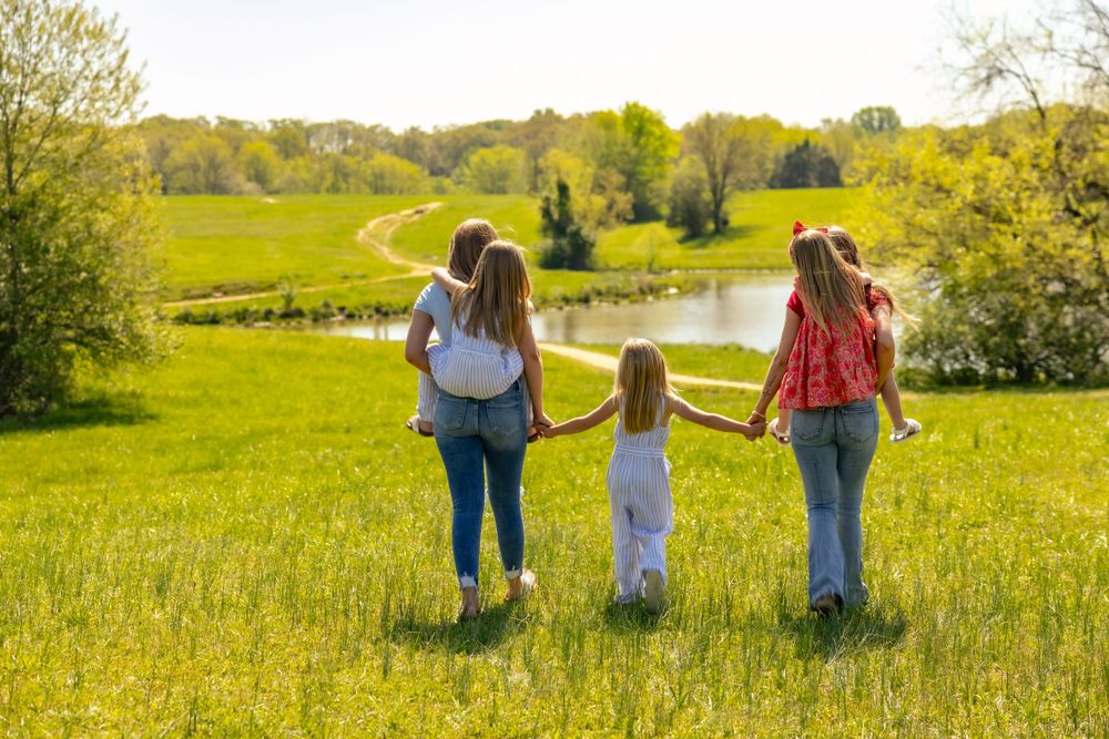 Three people walking hand in hand through a sunny meadow with rolling hills and a pond in the background.