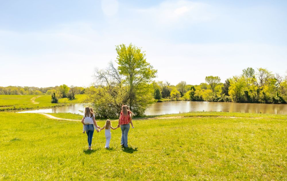 Three figures walk together on a rolling green hill with a scenic pond in the background.
