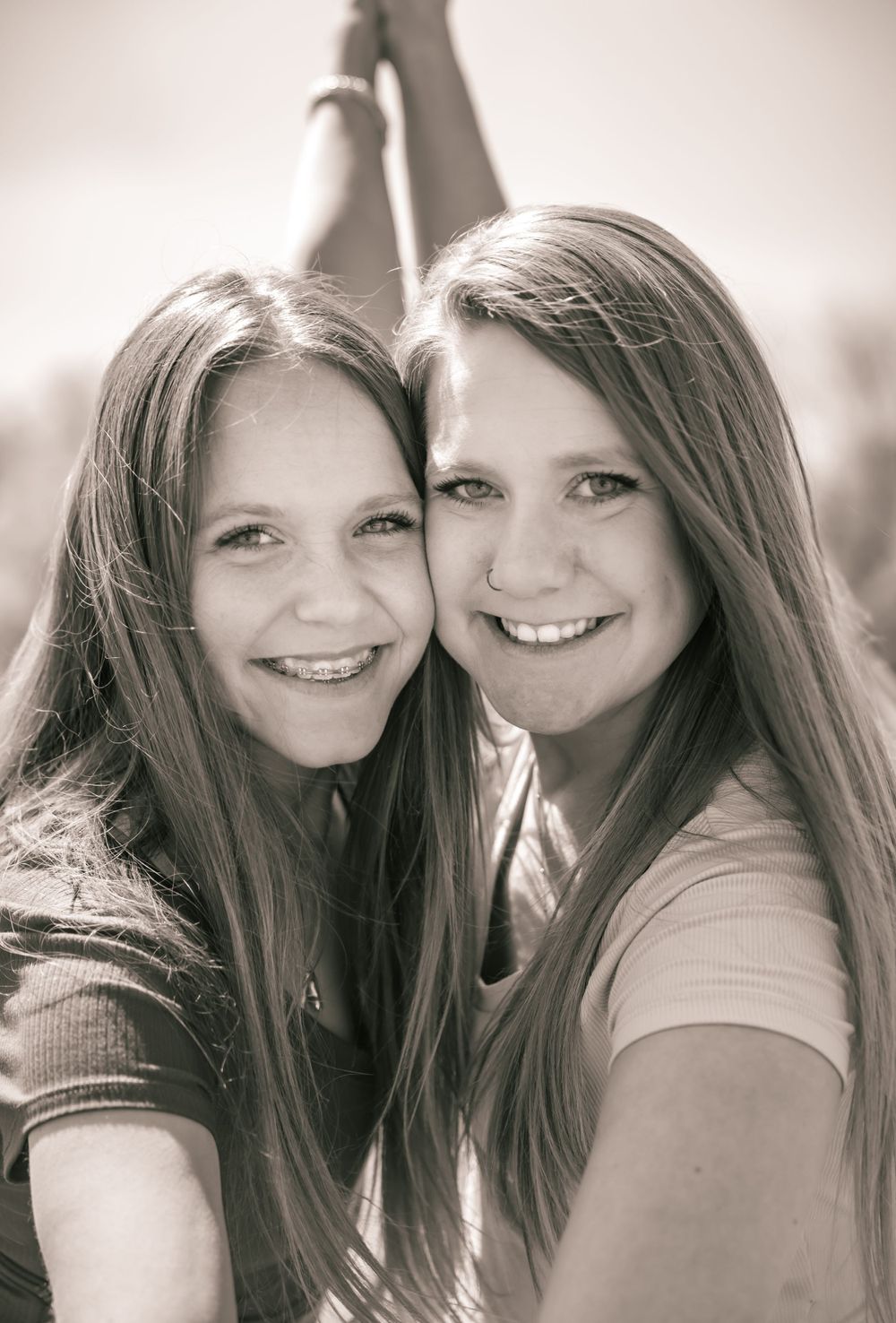 Two friends with long brown hair lean close together and smile warmly in a black and white portrait.