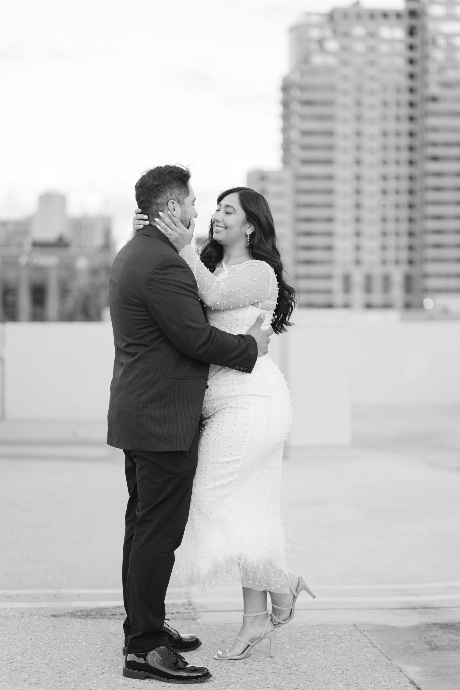 A black and white romantic wedding photo showing a tender embrace between a couple on a rooftop with city buildings behind them.