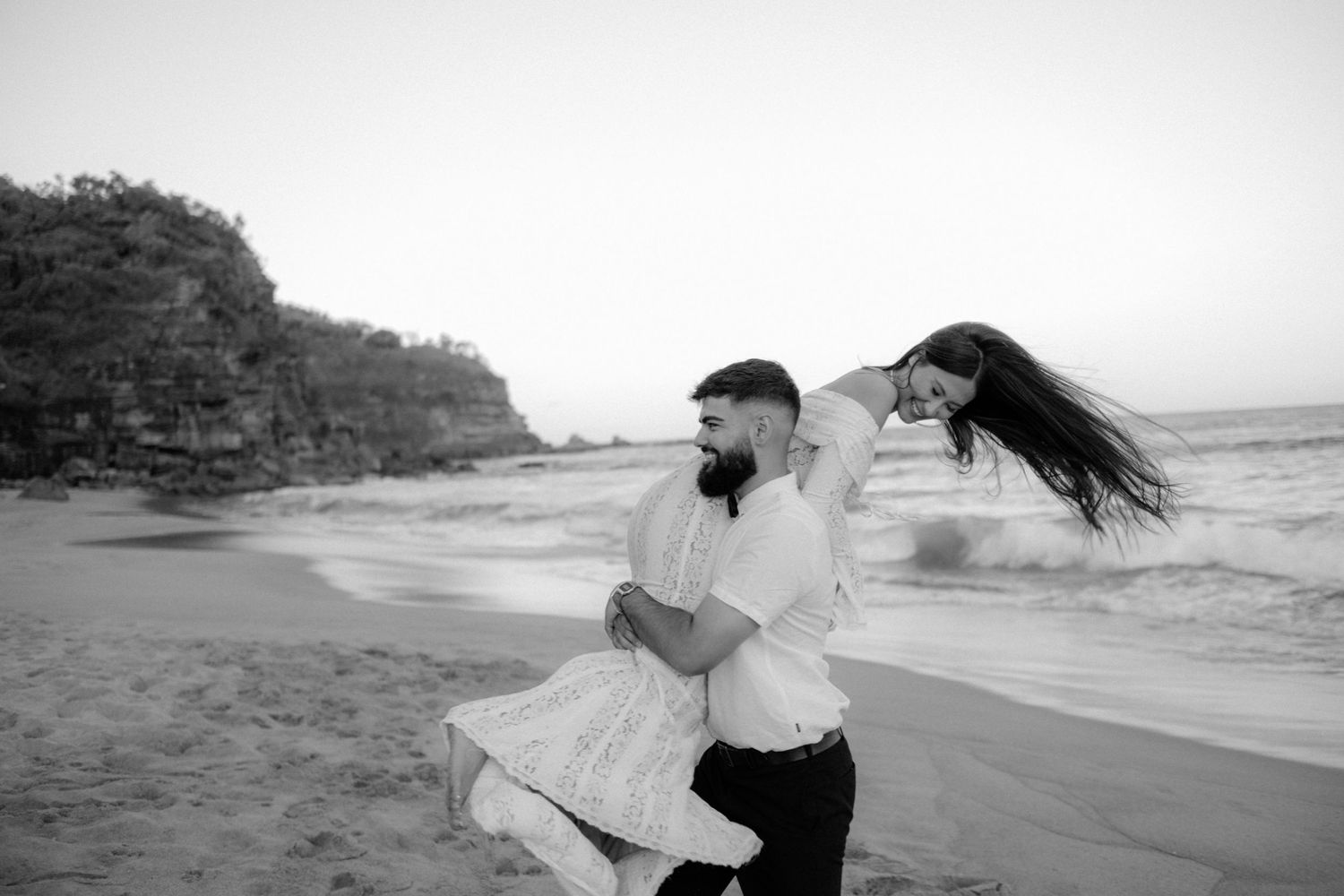 A couple in white clothing dances together on a beach at sunset with long hair flowing in the wind.
