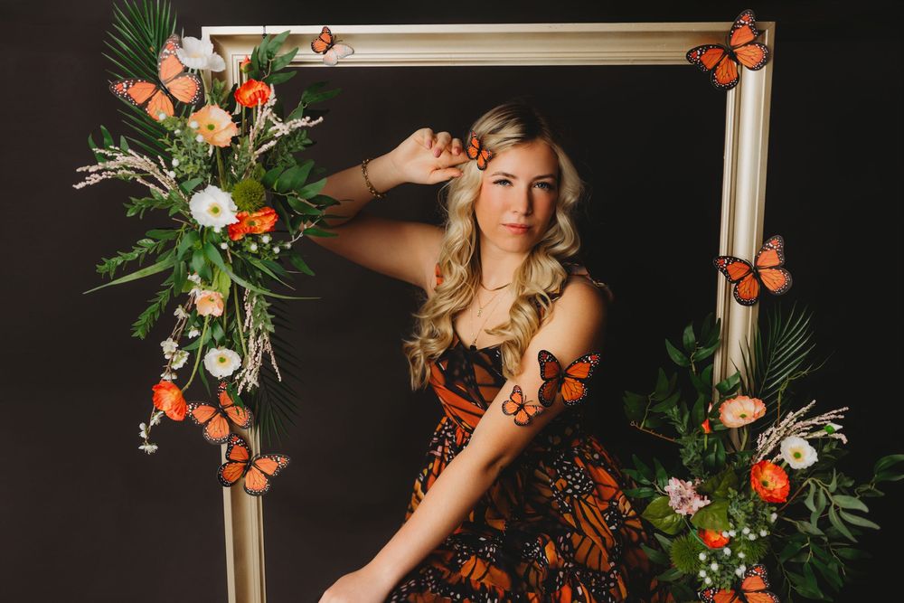 Artistic portrait with monarch butterflies and floral decorations against dark background.