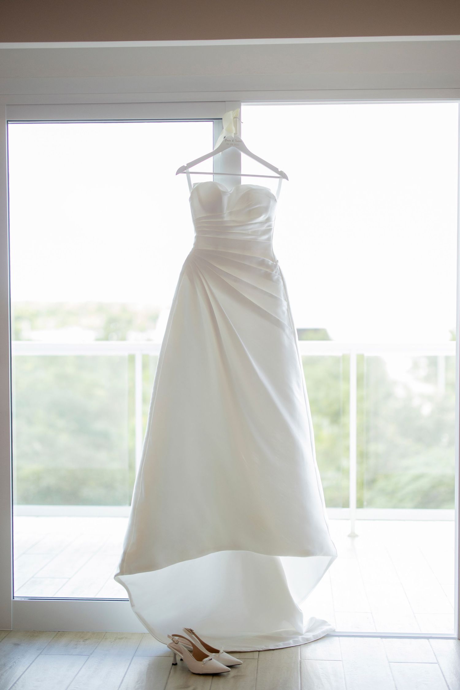 Elegant white wedding dress hanging by a window with bridal shoes underneath on a wooden floor in natural light.