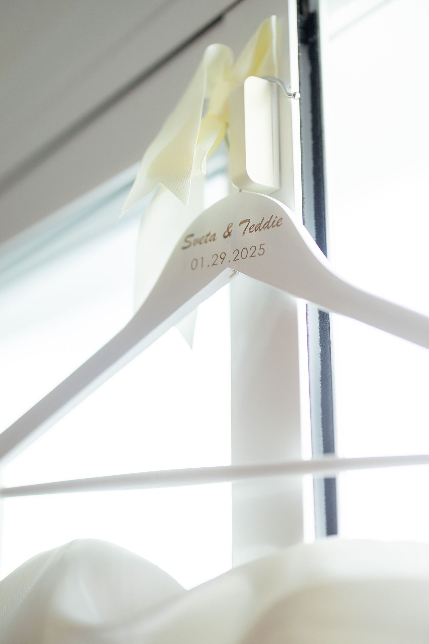 A white custom wedding hanger with names and date 01-29-2025 engraved, shown against a bright window.