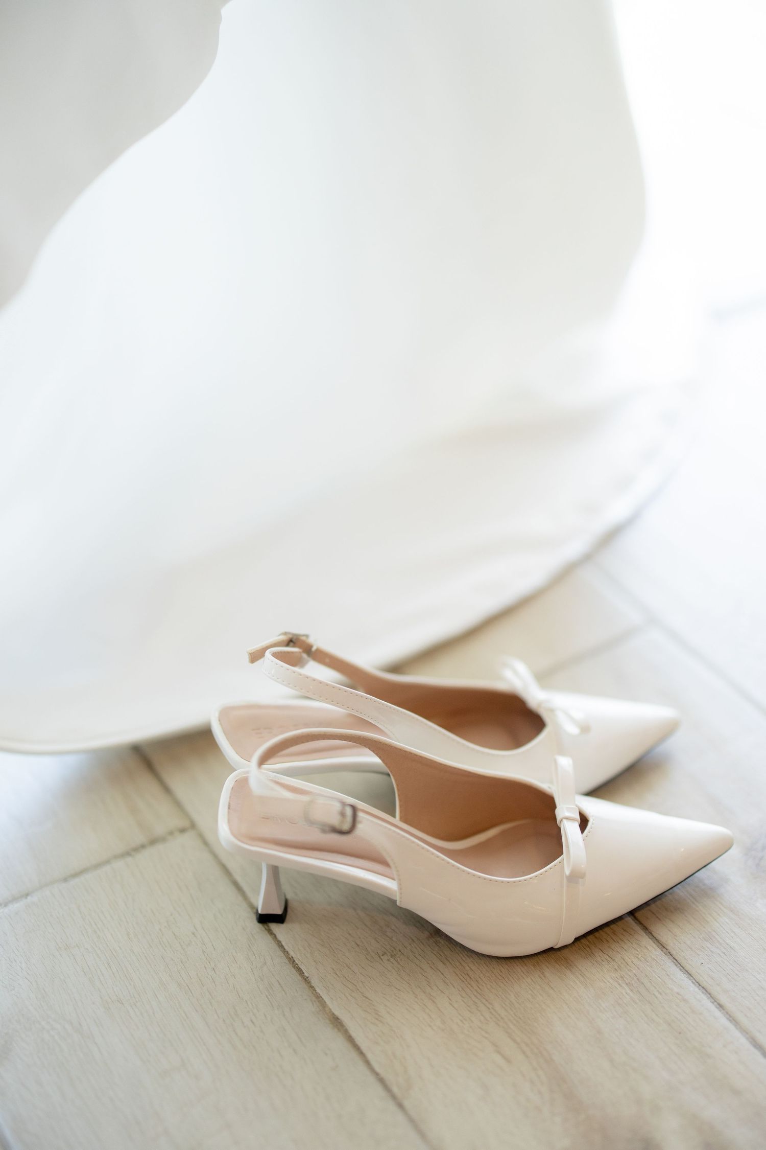 Elegant white slingback pumps with pointed toes and kitten heels sit on a light wooden floor.