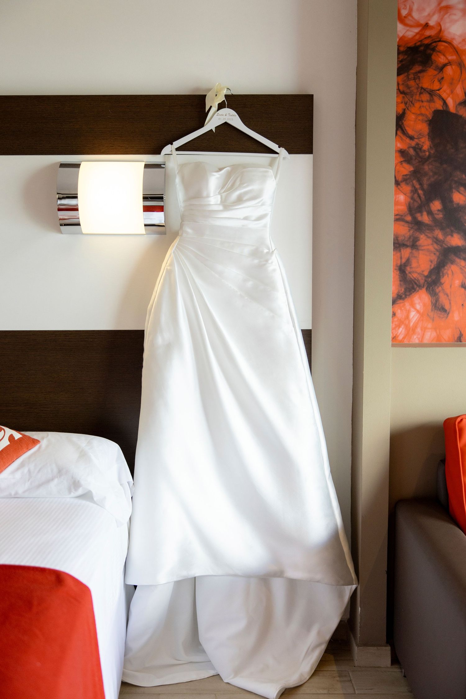 An elegant white wedding dress hanging on display in a modern hotel room with red and brown accents.