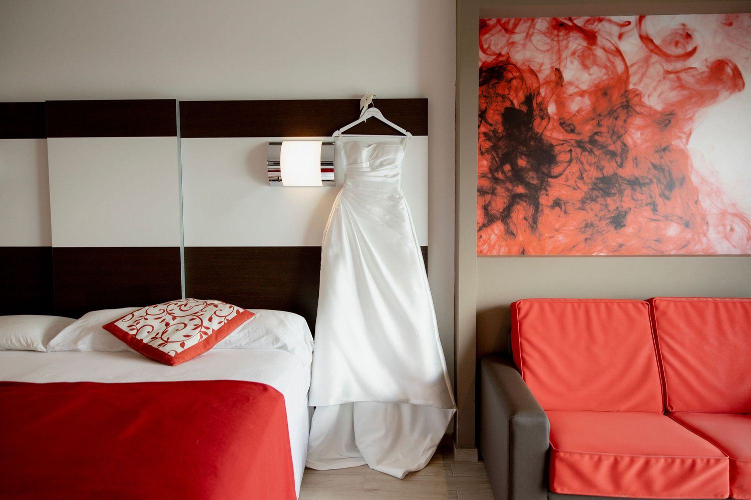 White wedding dress hangs above red bed next to coral sofa and abstract red wall art in modern bedroom.
