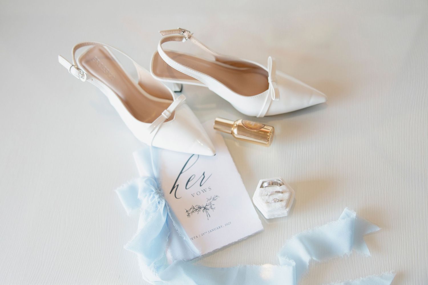 Wedding details with white pointed-toe bridal shoes, vow booklet, and delicate light blue silk ribbon on white surface.