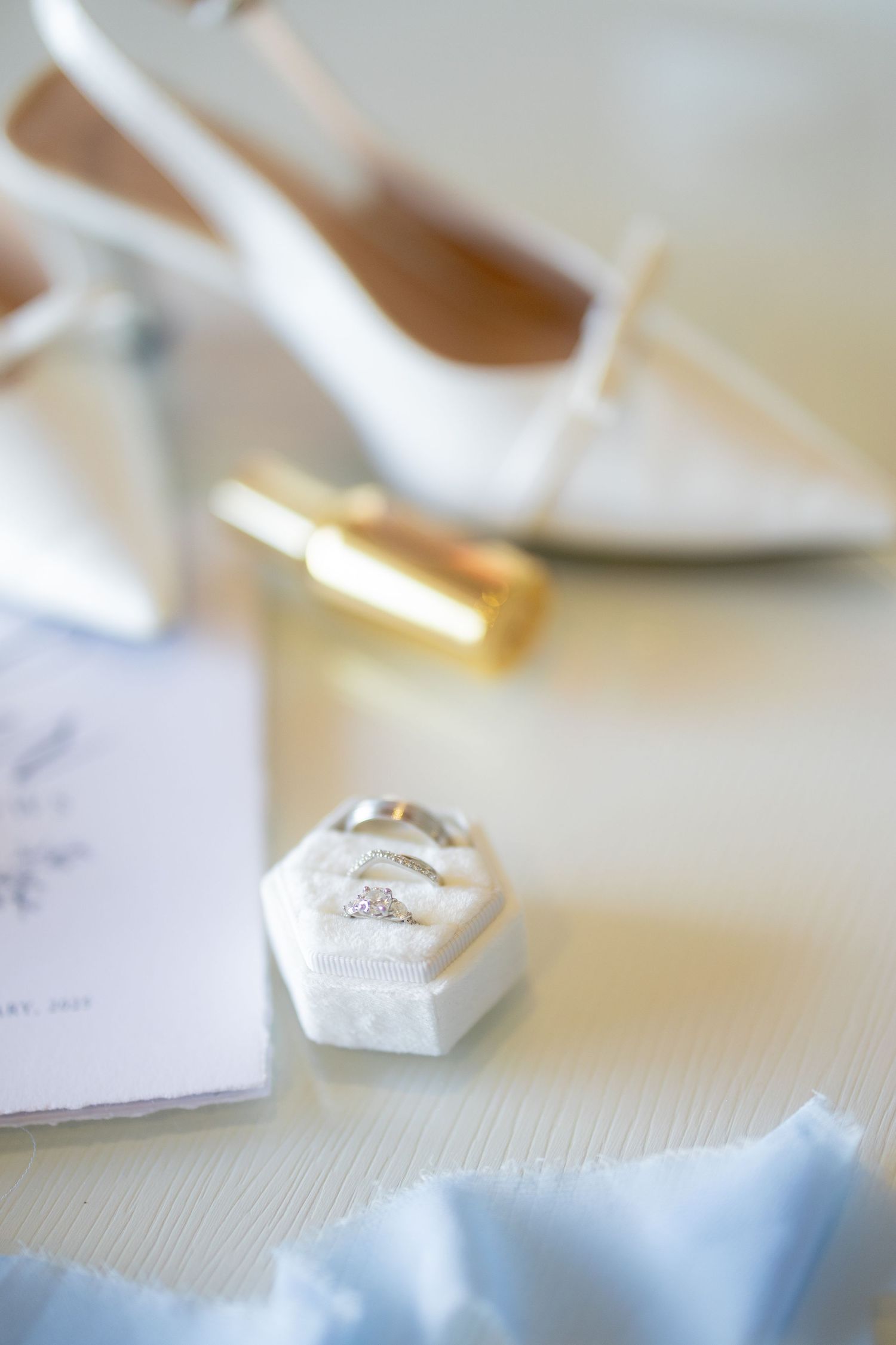 A delicate white wedding shoe and engagement ring box arranged with subtle blue decor on a light surface.