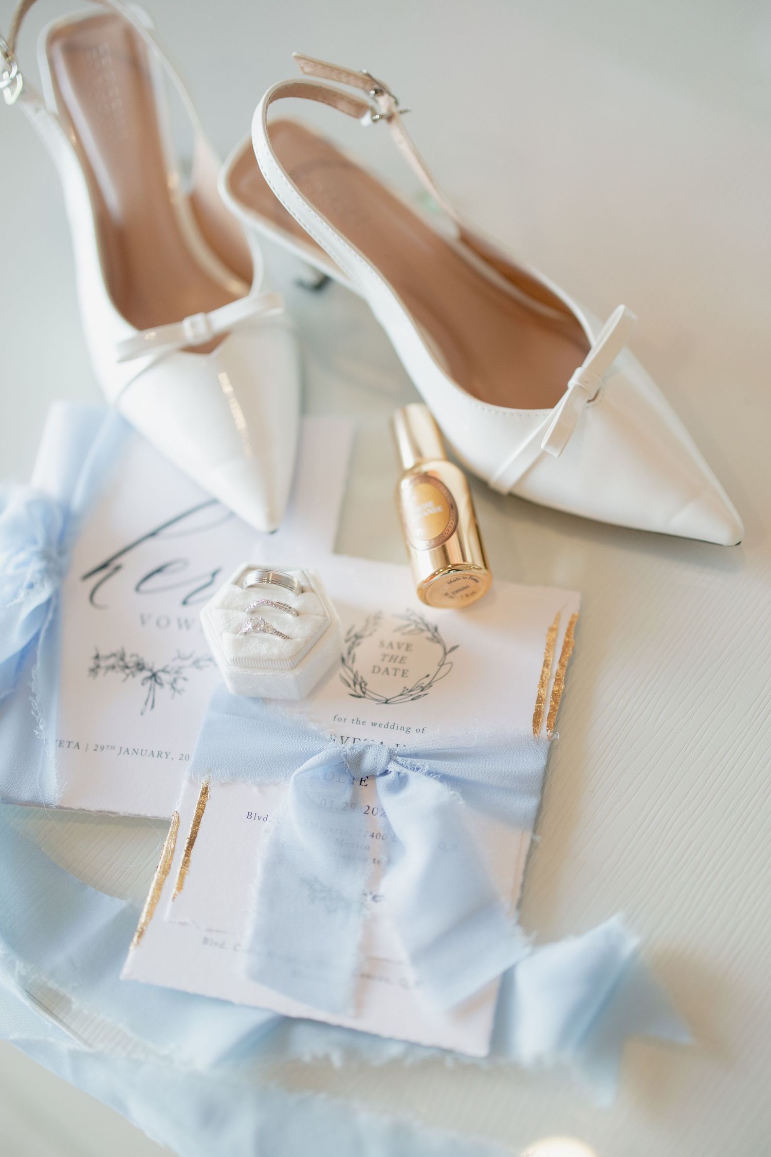 Elegant white wedding shoes with pointed toes and delicate straps styled with light blue ribbons and invitation details.