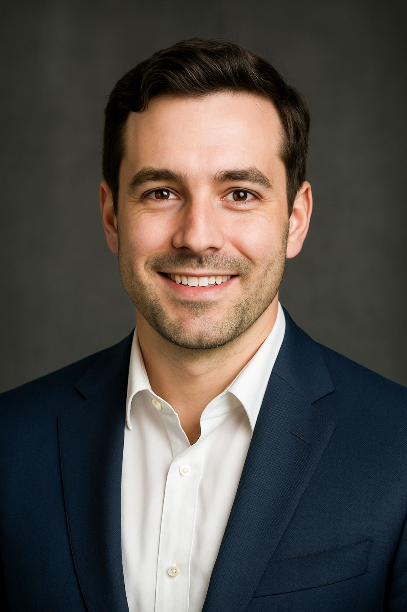 Professional headshot of a smiling person wearing a navy suit jacket and white button-down shirt against a dark background.