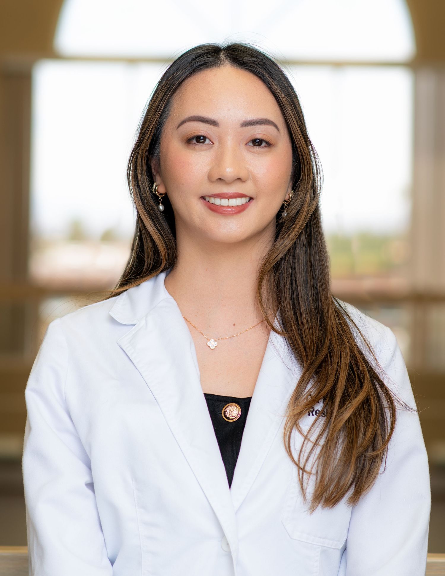 Healthcare professional in white lab coat and black top stands in medical office setting.
