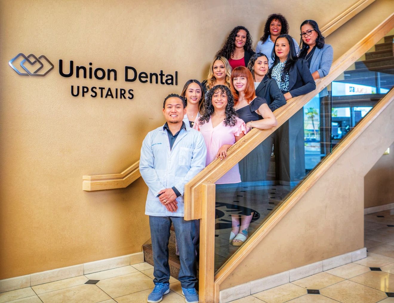A dental office team poses together on wooden stairs next to a Union Dental Upstairs sign on a gold wall.