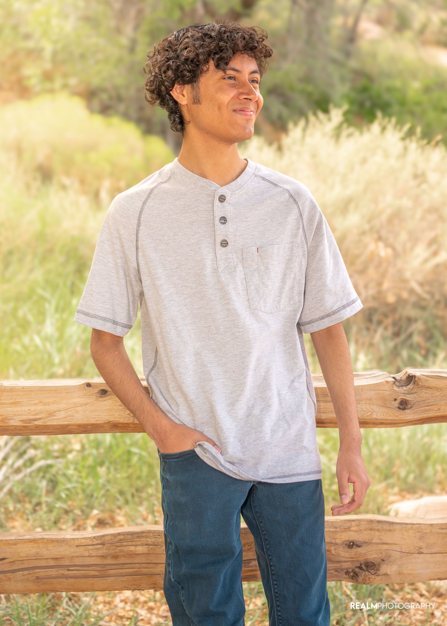 A person in a casual gray henley shirt and jeans smiles while leaning against a wooden fence in a sunlit outdoor setting.