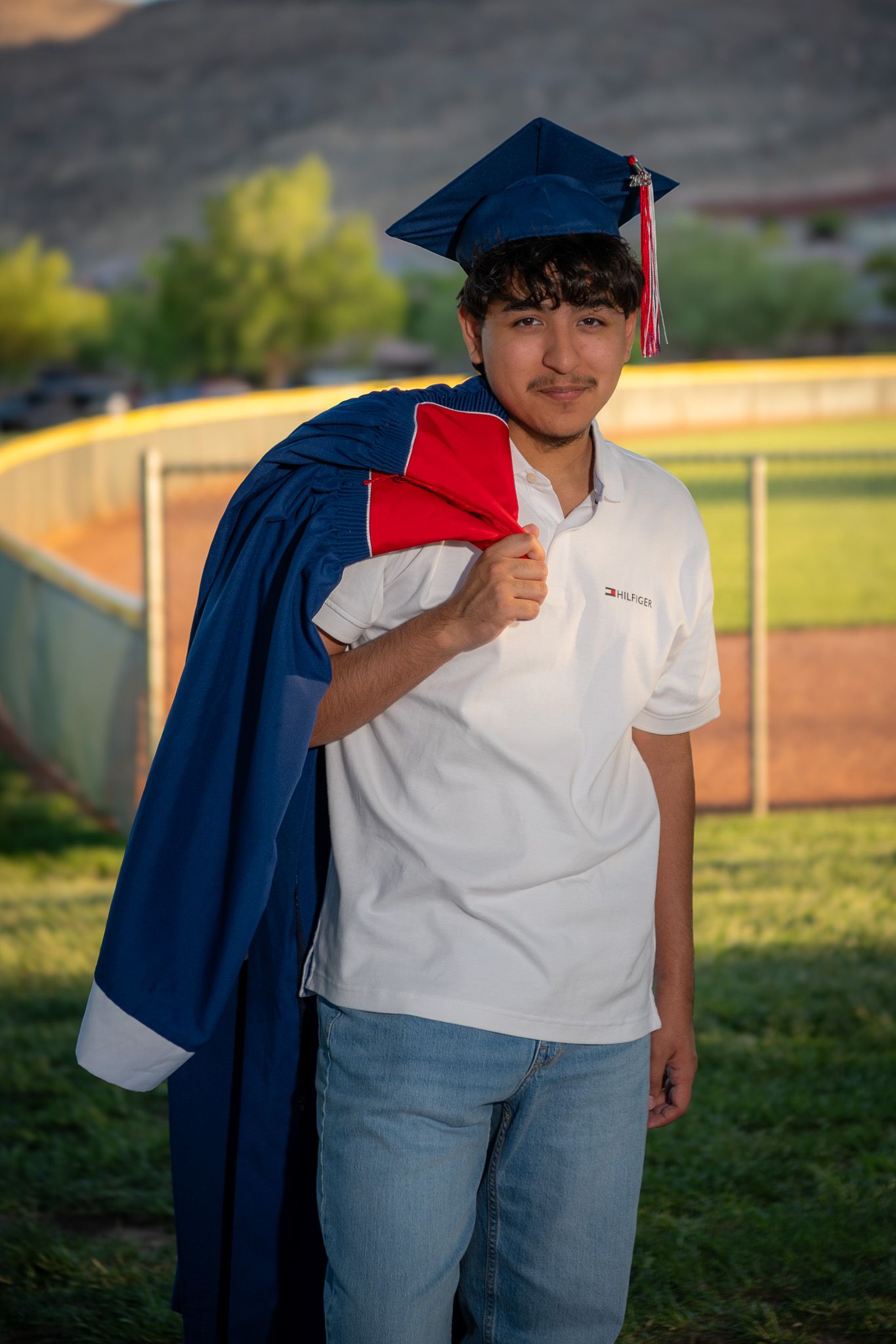 Graduate in blue cap and gown poses outdoors with mountains in background.