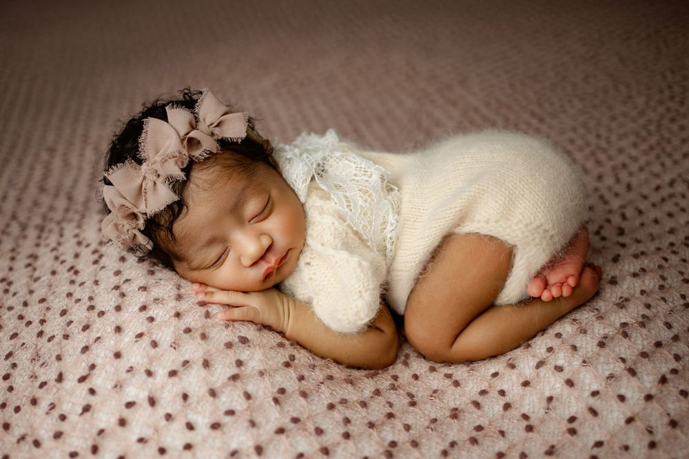 Sleeping newborn wearing white lace outfit and floral crown rests peacefully on textured pink blanket.