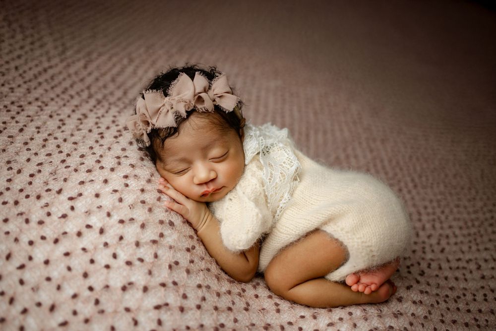 Sleeping newborn wearing white knit wrap and floral crown rests peacefully on textured mauve blanket.