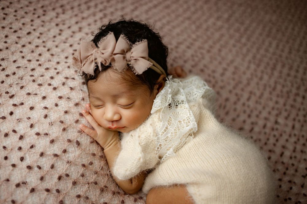 Sleeping newborn wearing white knit outfit and floral headband rests on textured pink blanket.