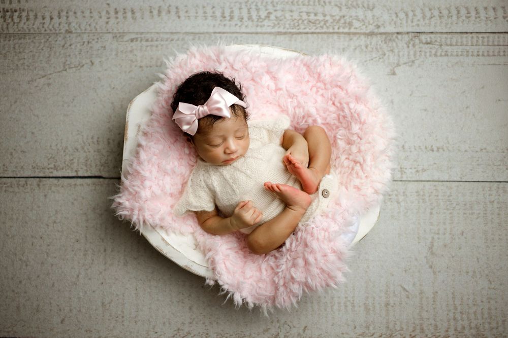 Sleeping newborn in white romper resting on fluffy pink blanket in studio setting.