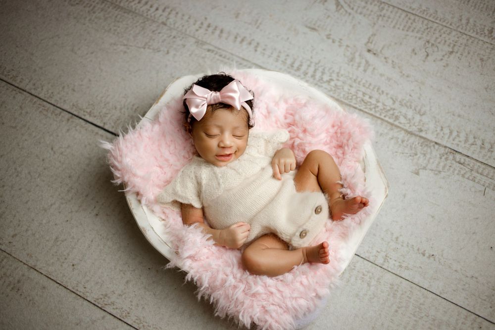 Infant wearing white onesie rests on pink fluffy blanket in white basket on wooden floor.