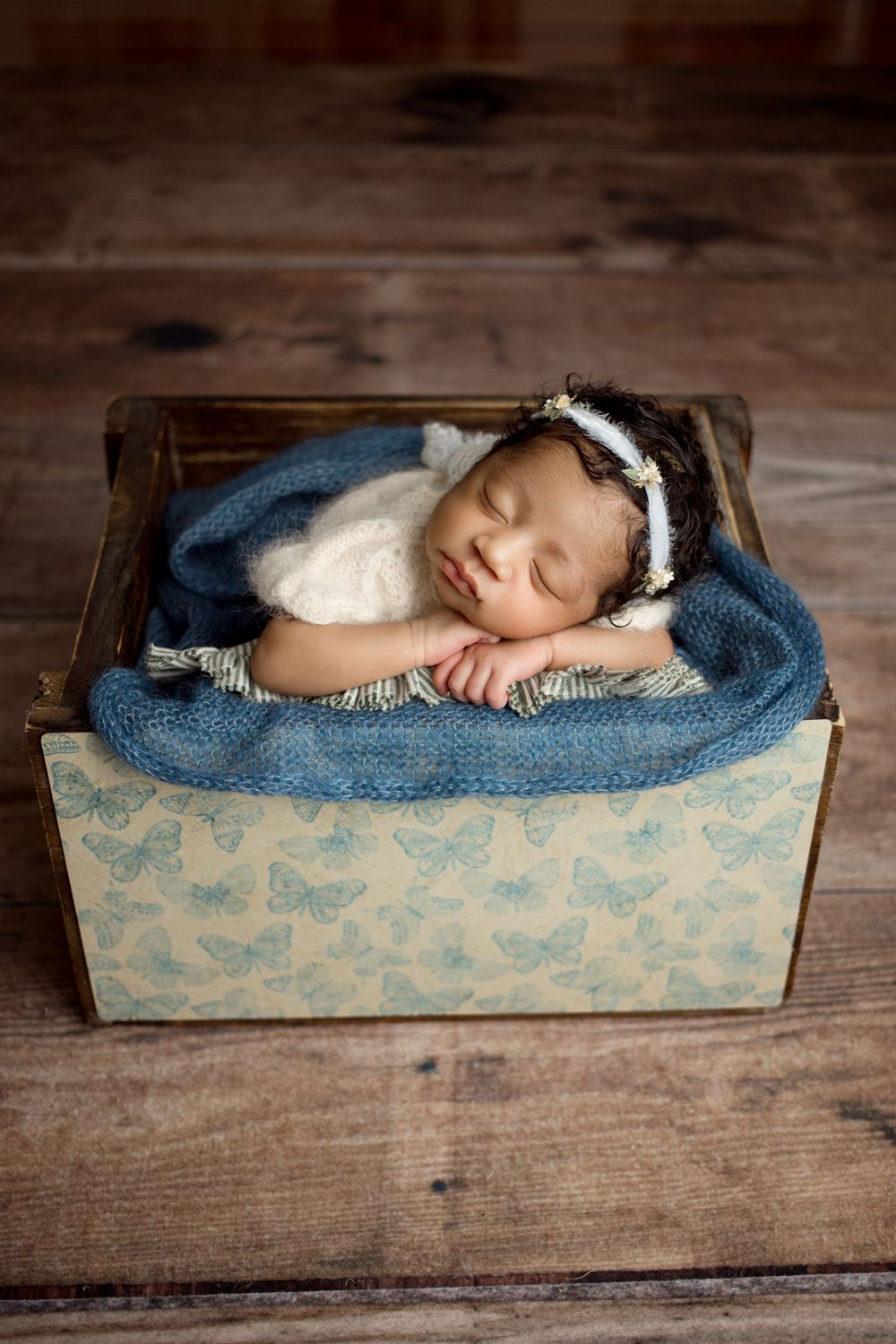 A sleeping newborn swaddled in blue blankets rests peacefully in a decorative wooden box on rustic floorboards.