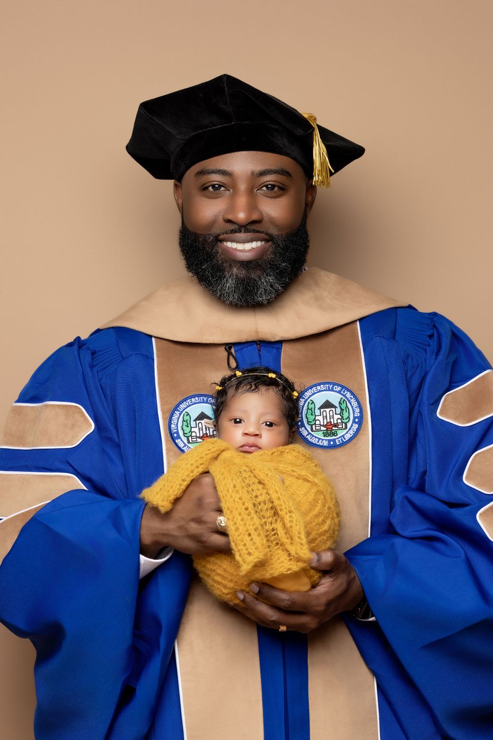 Graduate in blue and tan doctoral regalia holds small baby wearing yellow knit sweater during graduation photo.