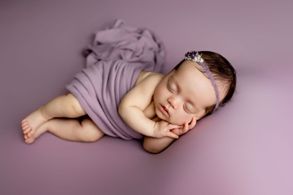 Peaceful sleeping newborn baby wrapped in soft purple fabric resting on matching purple background.