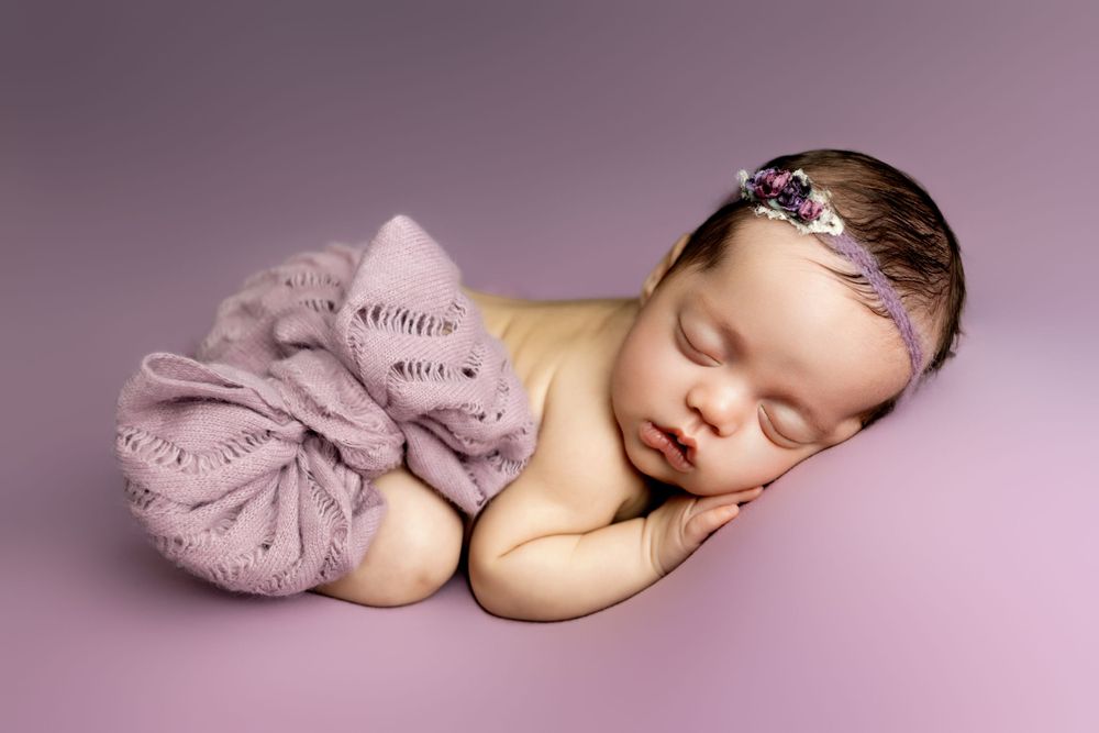 Sweet newborn baby sleeping on a lilac backdrop wrapped in soft purple fabric with delicate headband.