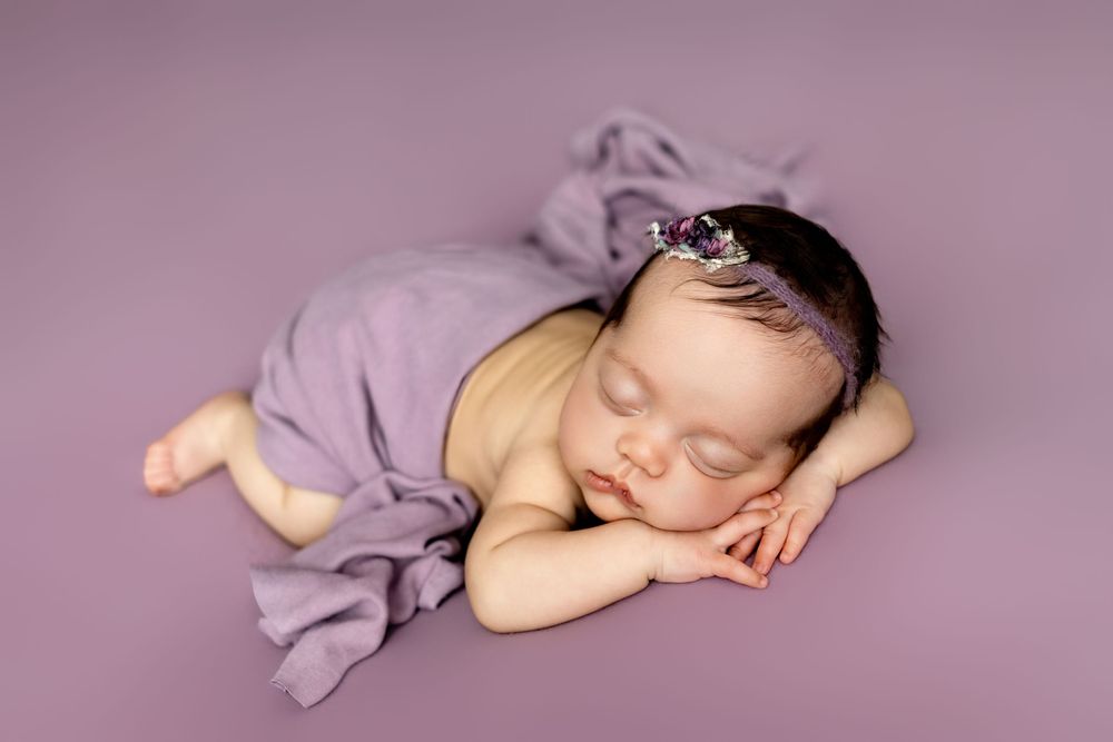 Sleeping newborn wearing purple wrap and tiny crown rests peacefully on purple backdrop during professional photo session.